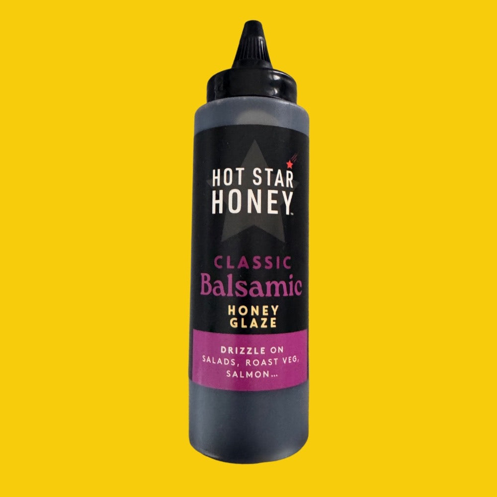 A squeeze bottle of Hot Star Honey Classic Balsamic Honey Glaze centered on a solid bright yellow background. The bottle is dark gray/black with a black label featuring white and pink text.