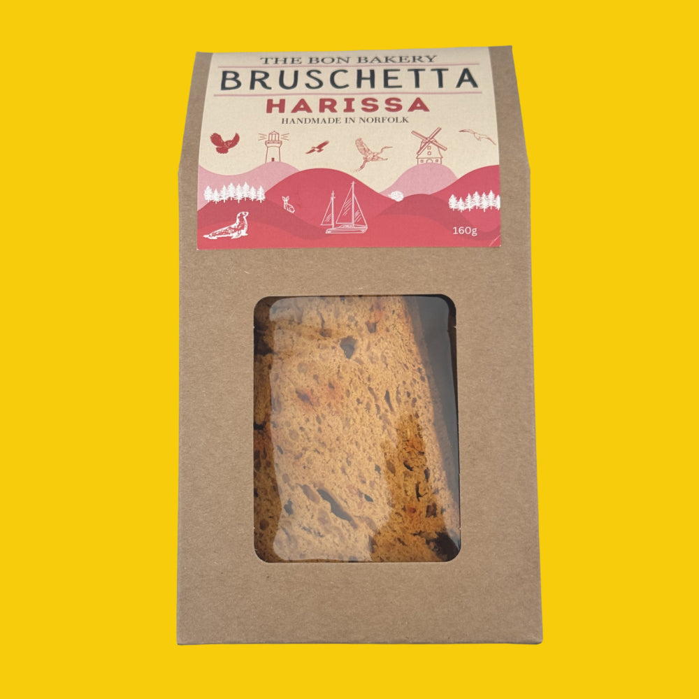 A package of The Bon Bakery Bruschetta Harissa handmade in Norfolk, shown in its brown kraft paper packaging with a clear window displaying the sliced harissa bread, set against a solid yellow background.