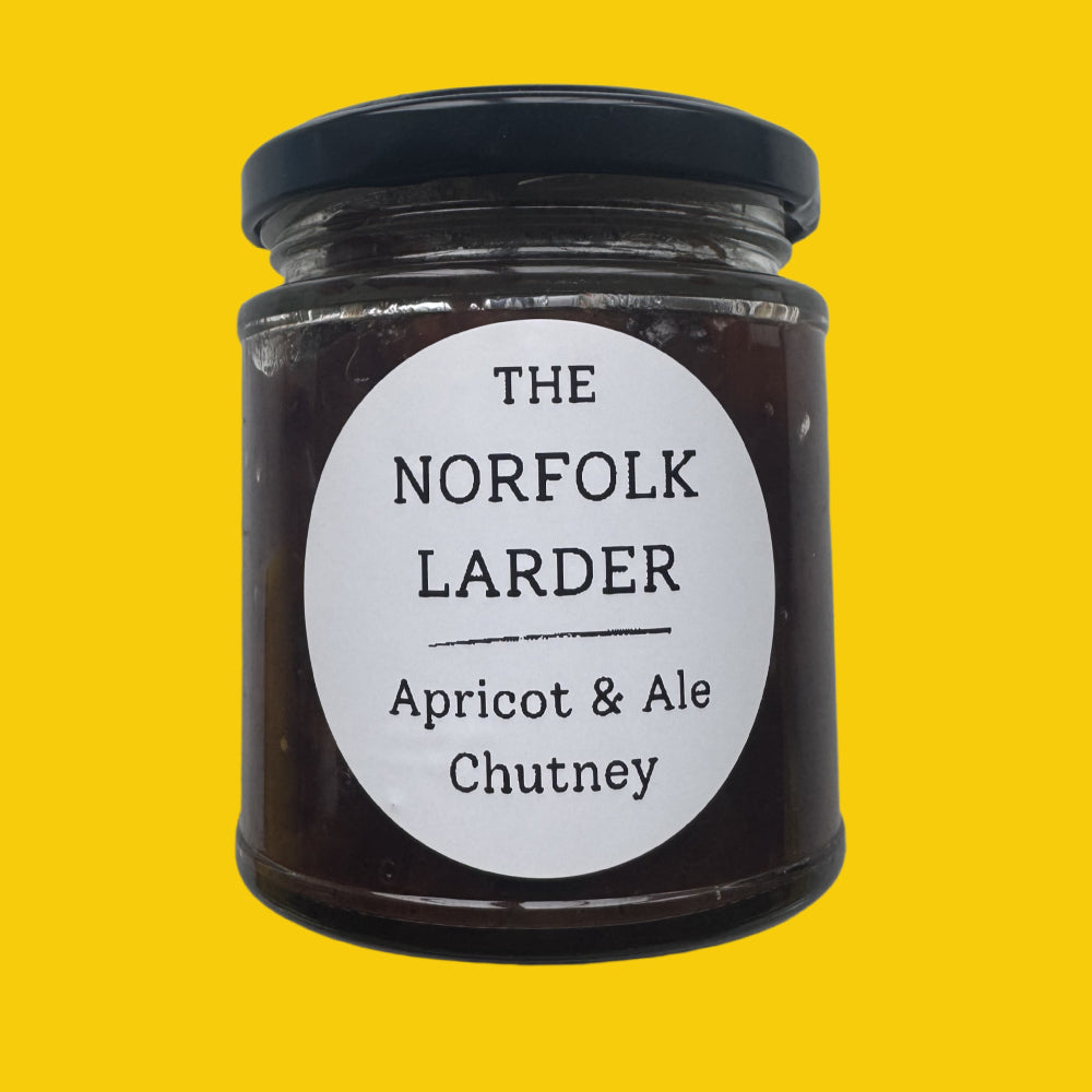 A glass jar of apricot and ale chutney with a black lid and a white circular label reading 'THE NORFOLK LARDER Apricot & Ale Chutney', centered on a solid yellow background.