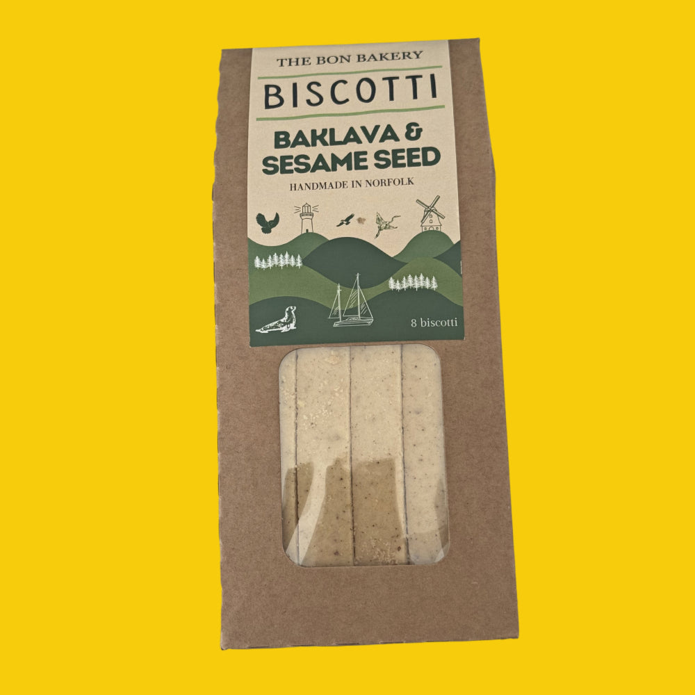 A package of Biscotti from The Bon Bakery, featuring Baklava & Sesame Seed. The product is in its brown kraft paper packaging with a clear window showing the baked biscotti inside, set against a solid bright yellow background.