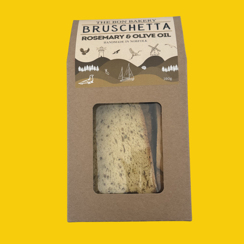 A 100g package of 'The Bon Bakery' Bruschetta Rosemary & Olive Oil handmade in Norfolk, shown in a brown kraft paper box with a clear window displaying the sliced bread and oil inside, set against a solid yellow background.
