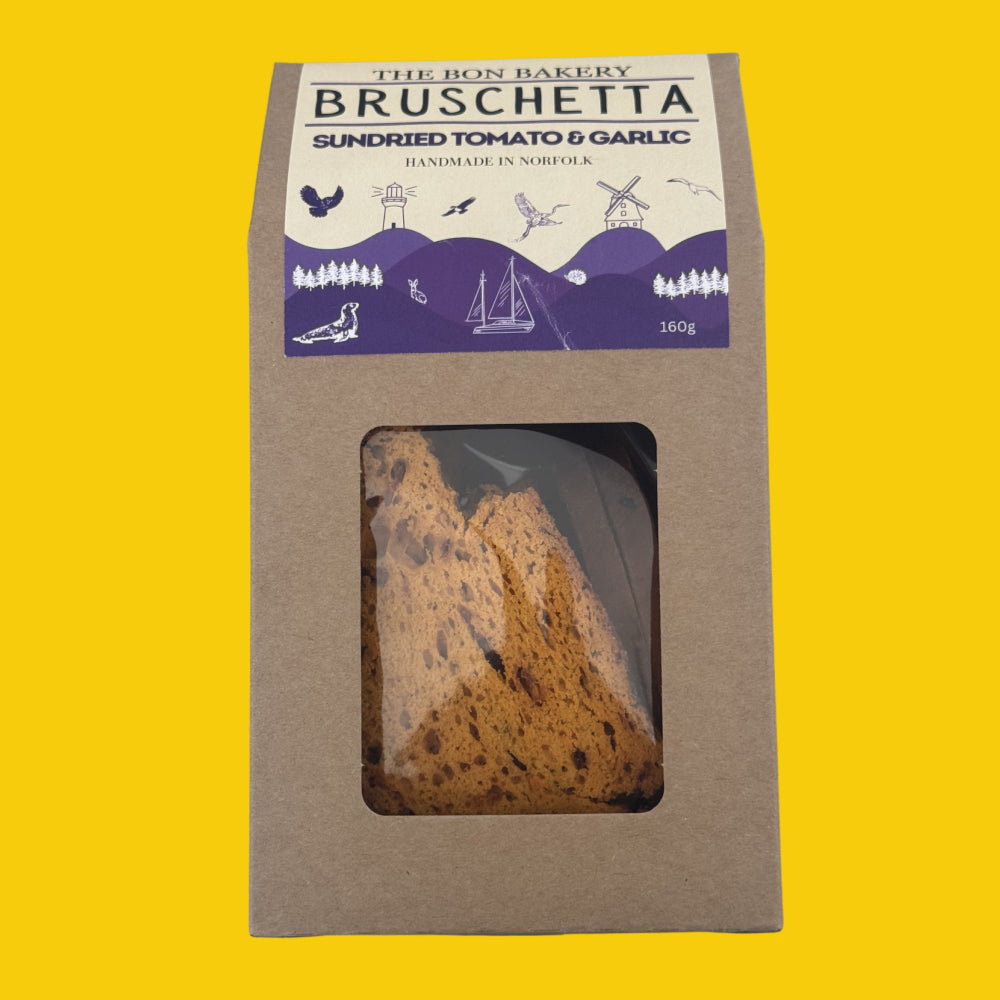 A package of The Bon Bakery Bruschetta Sundried Tomato & Garlic bread slices, shown in a brown kraft paper bag with a clear window displaying the golden-brown bread, set against a solid yellow background.